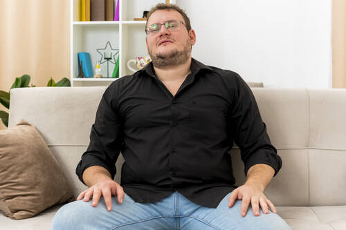 Confident adult slavic man optical glasses sits armchair putting hands legs inside living room