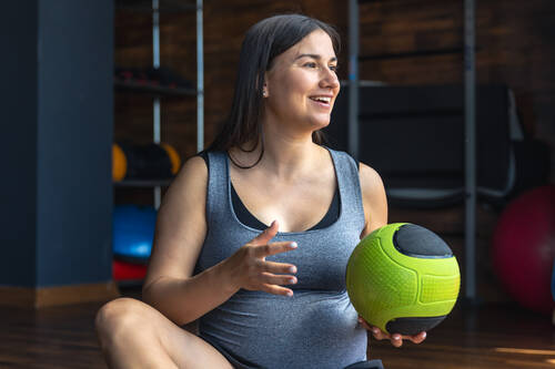Pregnant woman gym sits mat with ball her hands